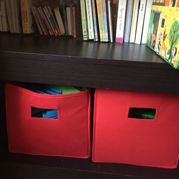 Storage bins in red on bottom shelf of bookshelf holding toys