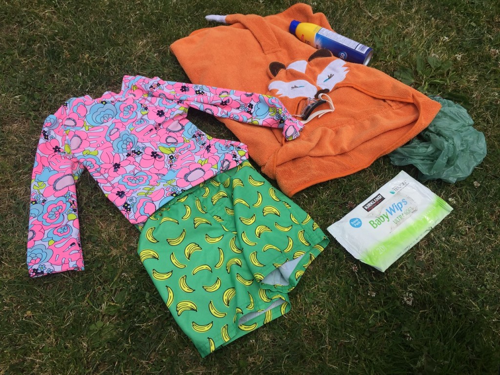 Swim shirt, swim shorts, sunscreen, hooded, towel, wipes, goggles, and plastic bag laying on grass