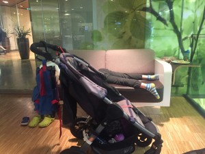 Empty stroller with bags handing off back handle with kid passed out on sofa in background