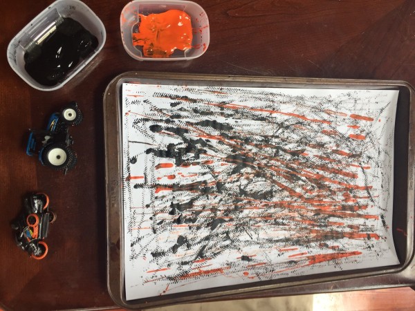 Paint by car supplies orange and black paint in small plastic bowls, paper on cookie sheet decorated by driving cars through paint and onto paper