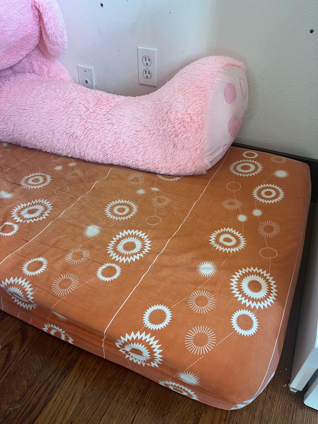 Simmons Kids SlumberTime Naturally 2 Stage Premium Plant Based Soy Foam Crib Mattress on floor in corner of room with orange pattern sheet and giant pink teddy bear